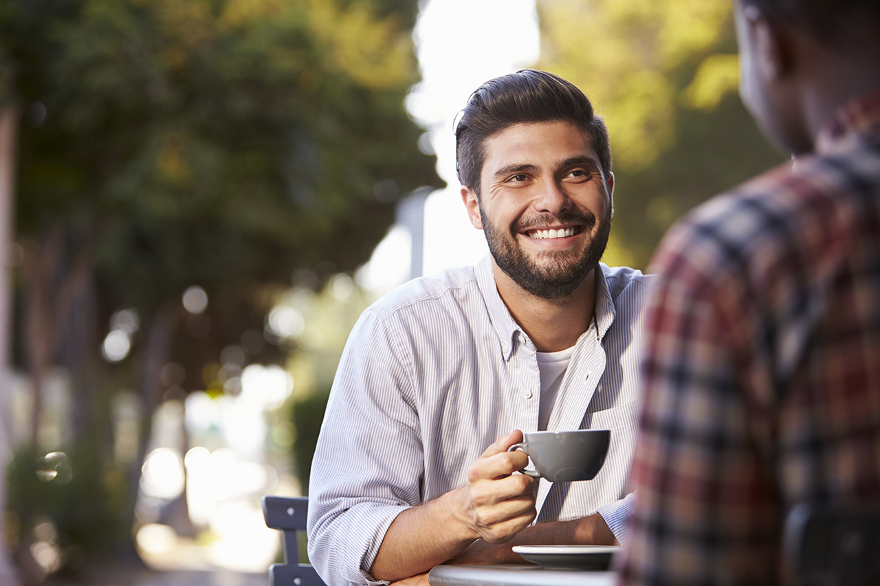 Two adult male friends sit talking over coffee outside cafe